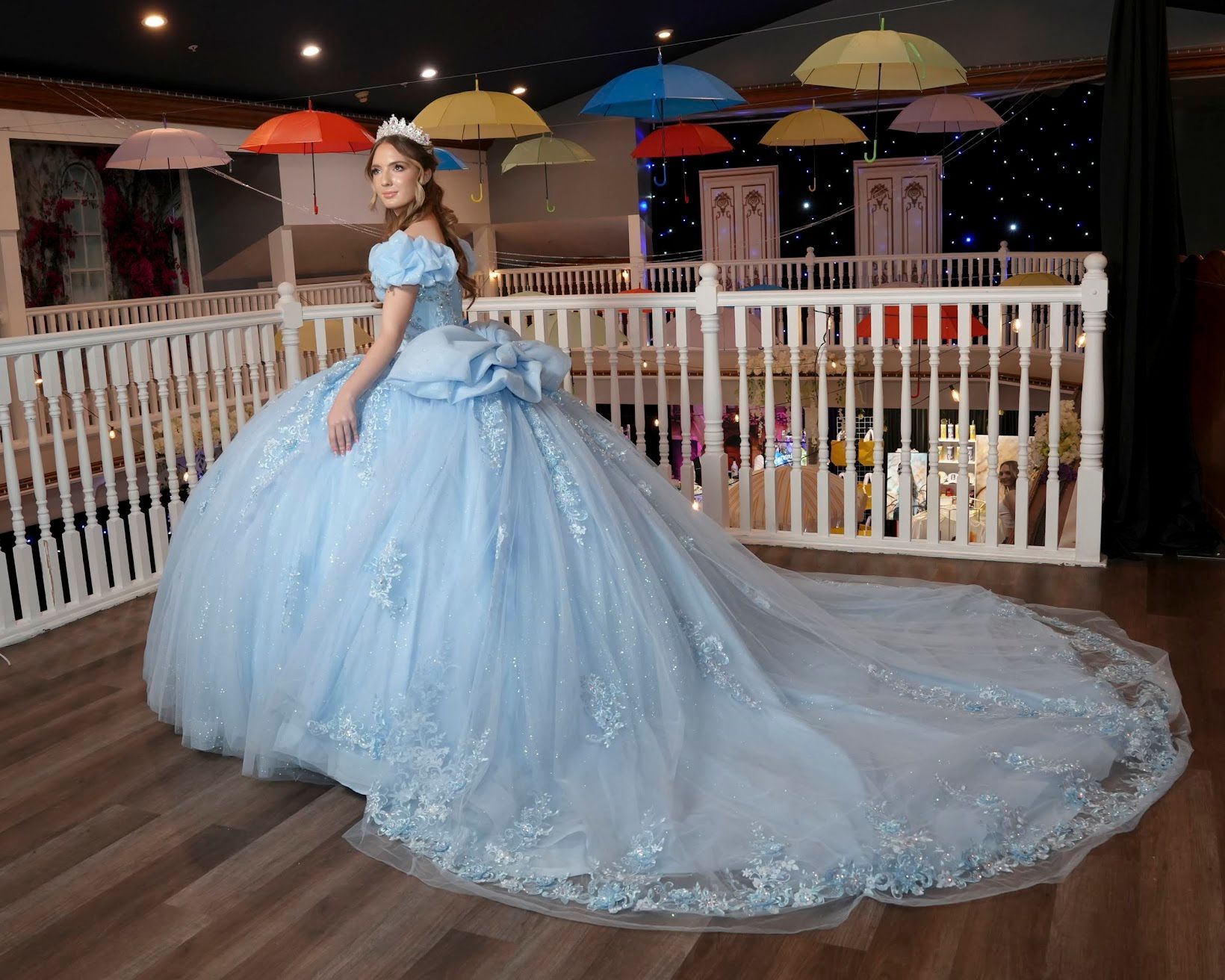Woman in a Cinderella-style blue gown with long train, posing on a balcony with umbrellas overhead.
