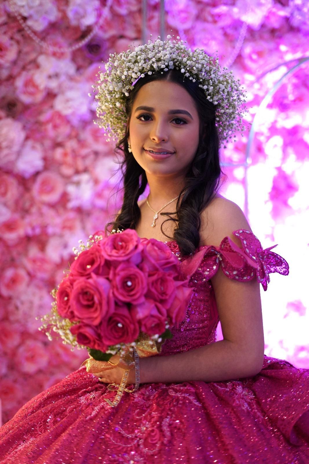 Woman in pink gown with flower crown, holding pink rose bouquet, in front of pink floral wall.