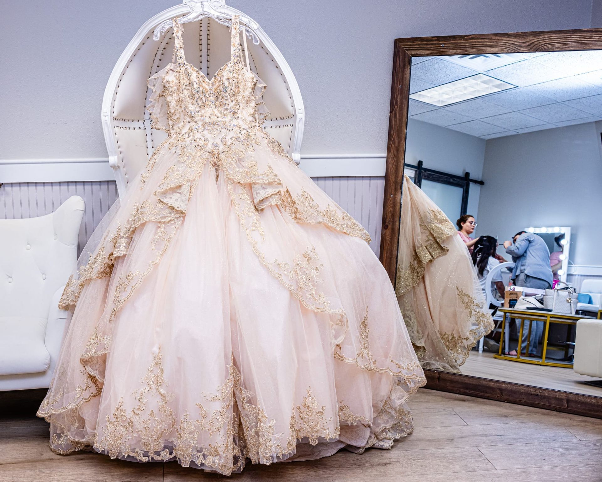 Elegant blush ball gown hanging in a white chair, gold embellishments, large mirror reflecting a person getting their hair done.