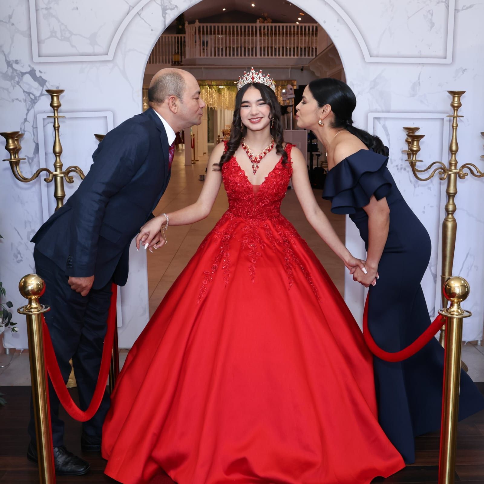 Woman in red gown with parents; kisses on cheeks, decorative arch, red carpet.