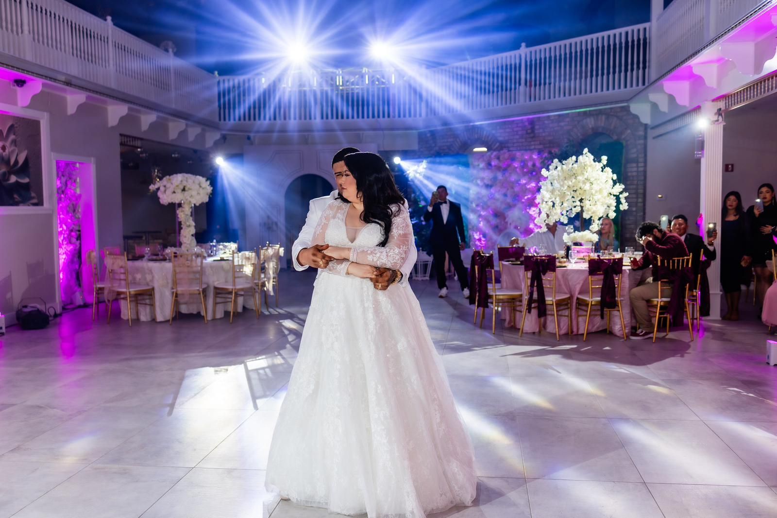 Couple dancing at a wedding reception, surrounded by tables, decorations, and stage lighting.
