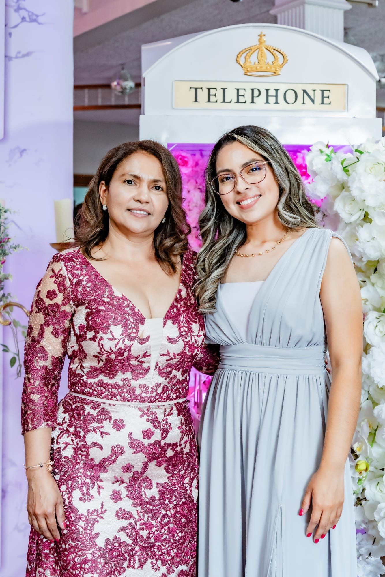 Two women posing in front of a white phone booth; one in a burgundy dress, the other in a gray gown.