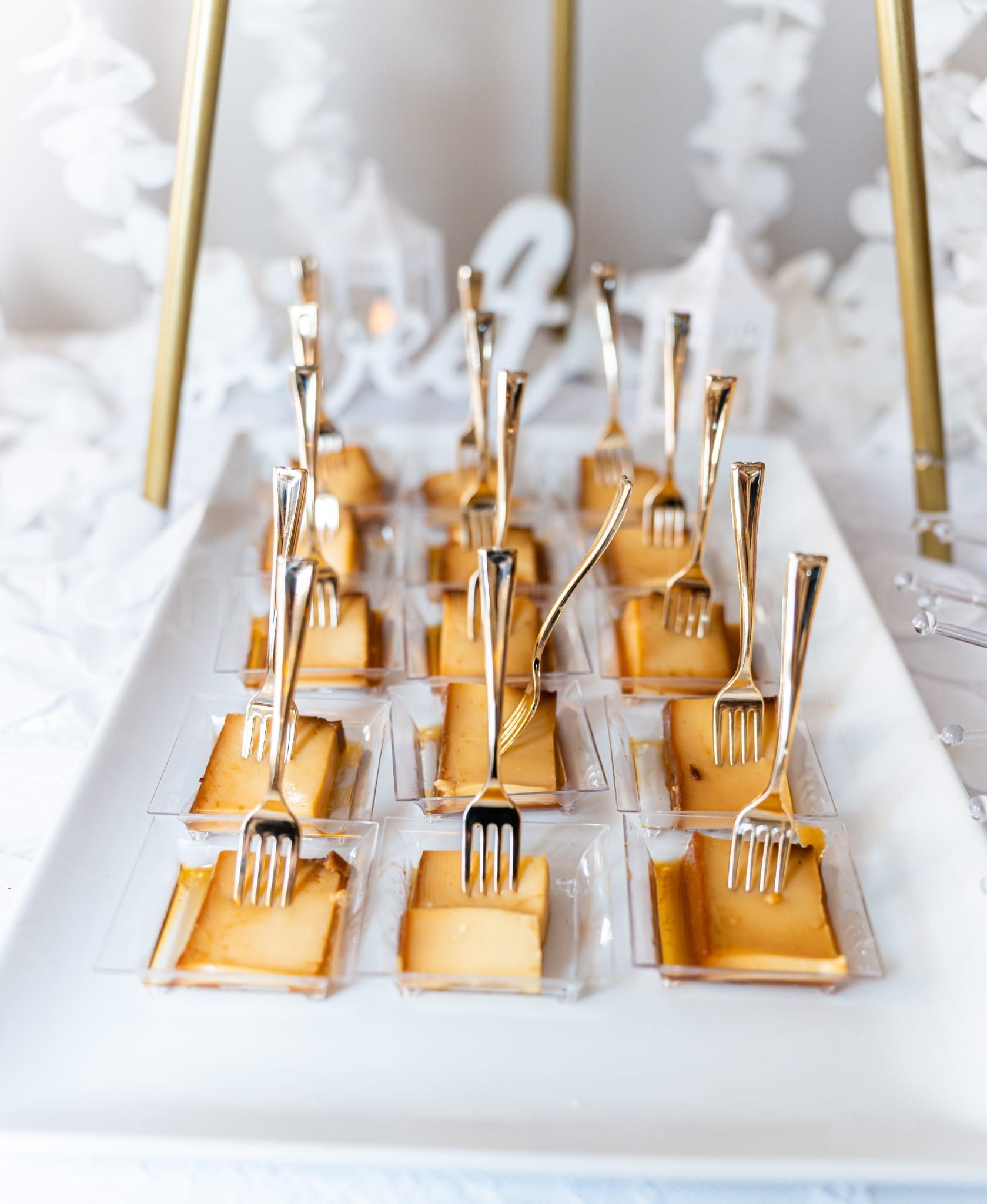 Appetizers on a white tray, each topped with a small gold fork. Gold frame in the background.