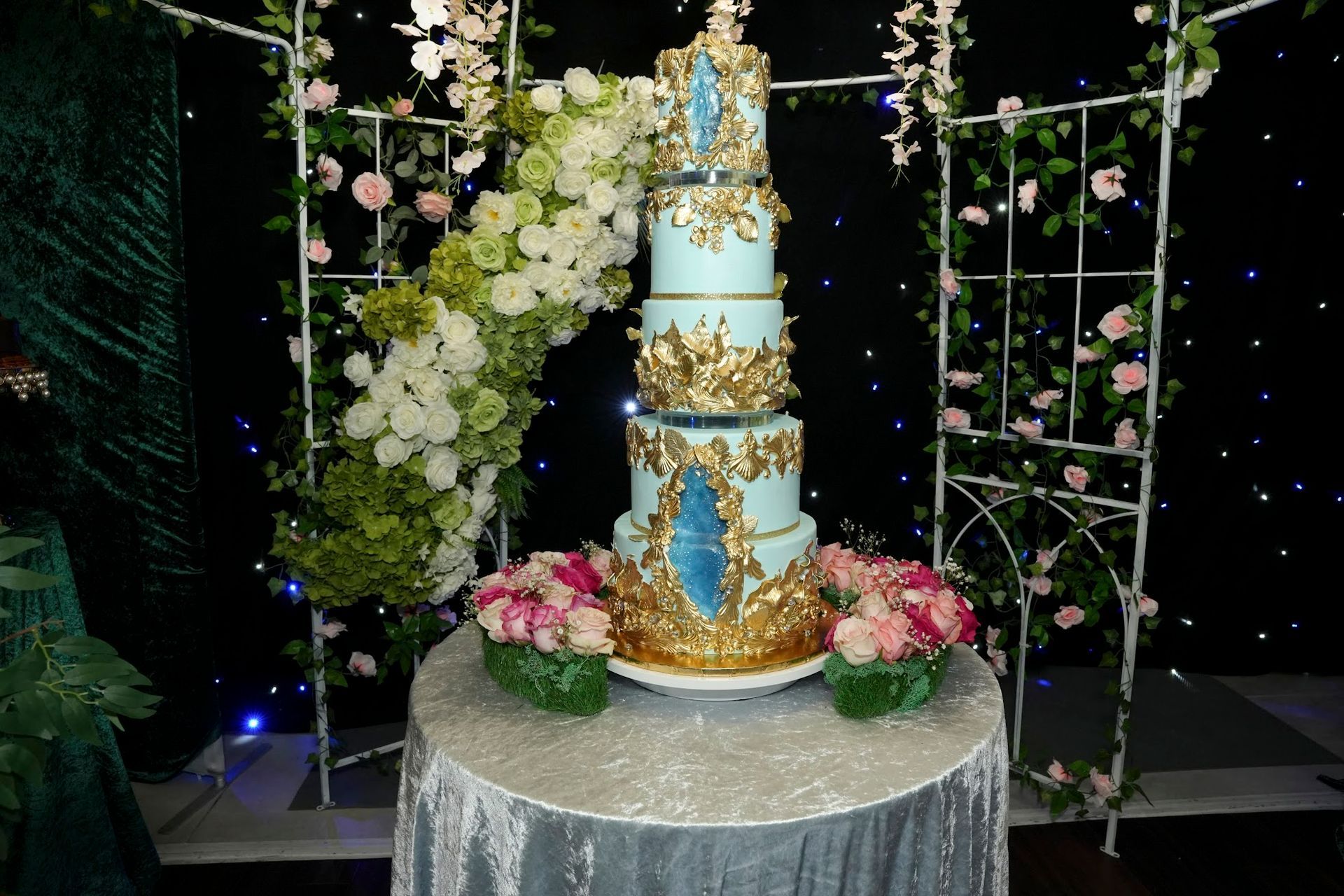 Five-tier blue cake with gold details, on a table. Floral arch and pink flowers surround it.