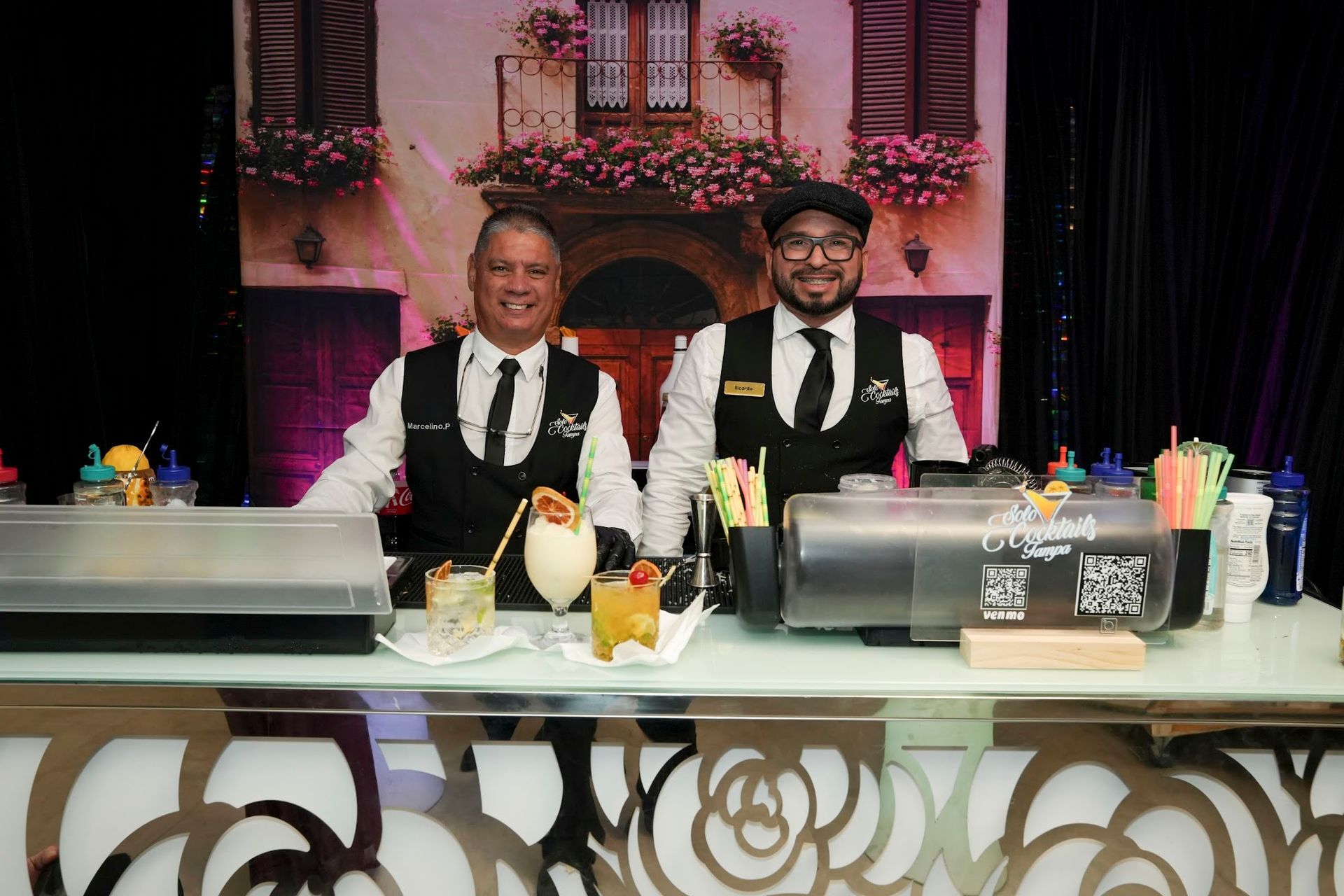 Two bartenders behind a bar serving cocktails; floral bar front, background scene, and colorful straws.