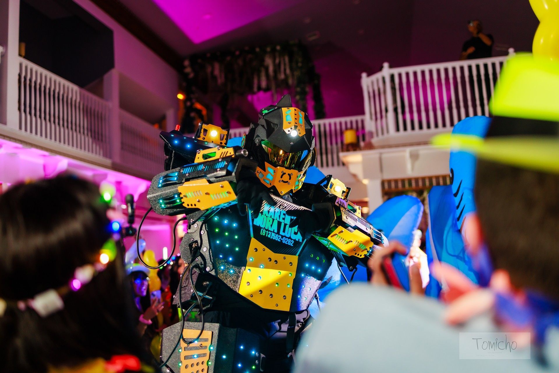 Robot performer in a neon-lit suit, interacting with a crowd indoors, at a brightly lit event.