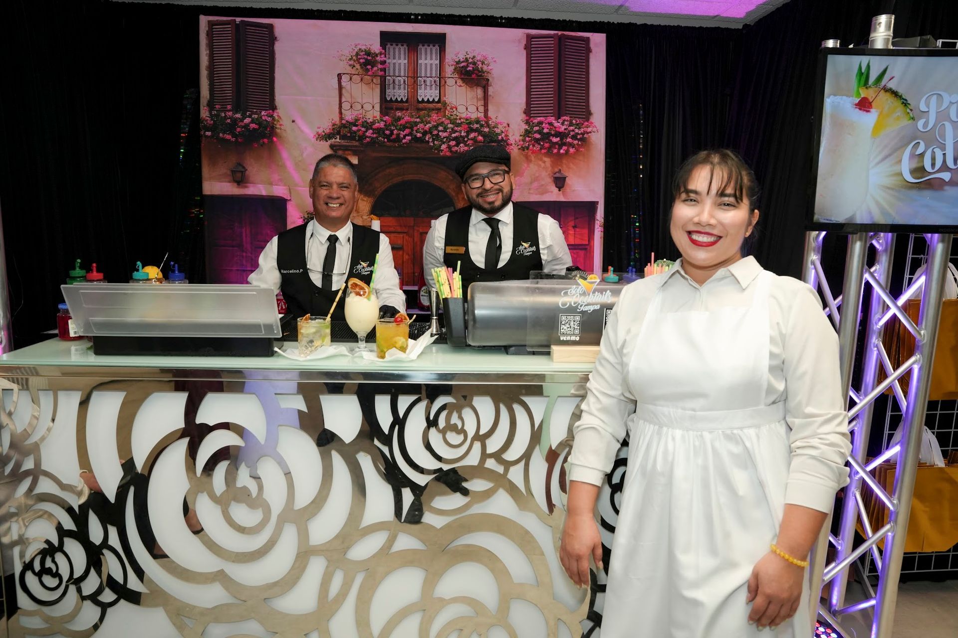 Three people behind a decorated bar, preparing drinks. A woman in a white dress smiles.  Two men wear vests and bowties.