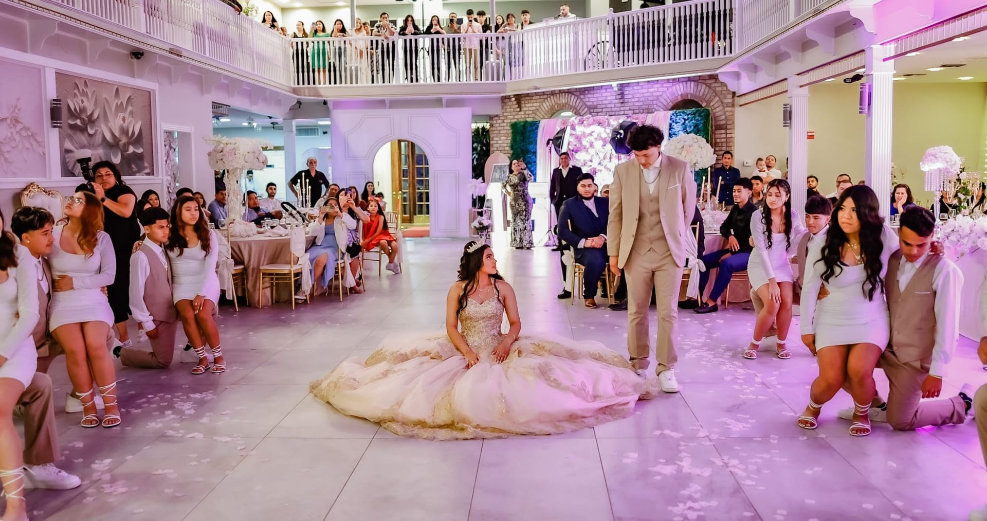 Quinceañera celebration. Young person in gown, central to group kneeling on dance floor. Guests seated, balcony above.