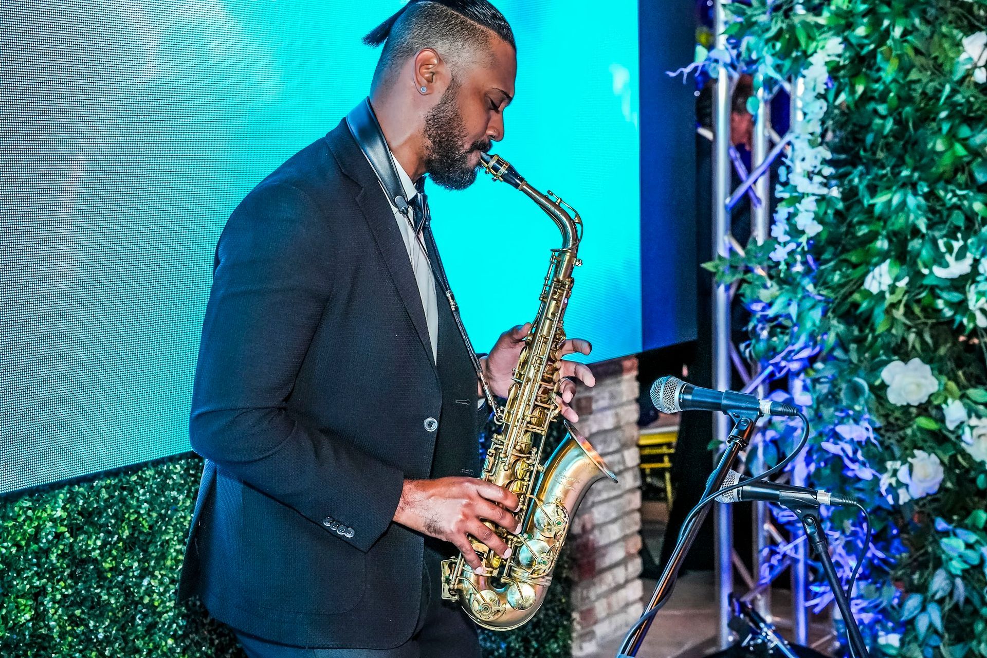 Man playing a saxophone at an event. He wears a dark suit, stands near a mic, and a green floral backdrop.