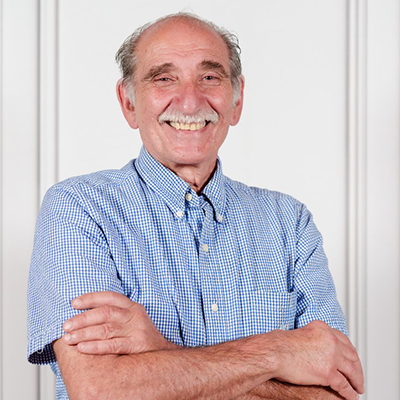 Man with mustache smiles, arms crossed, wearing a blue checkered short-sleeved shirt.