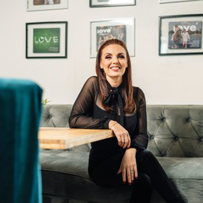 Woman with red hair, smiling, seated at a table in front of a gray couch, black top and pants, framed art on the wall.