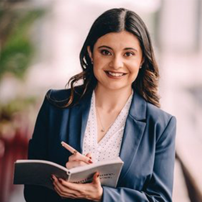 Woman in a blue blazer and white blouse, smiling and writing in a notebook.