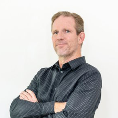 Man with arms crossed, wearing a dark button-down shirt, smiles slightly against a white background.