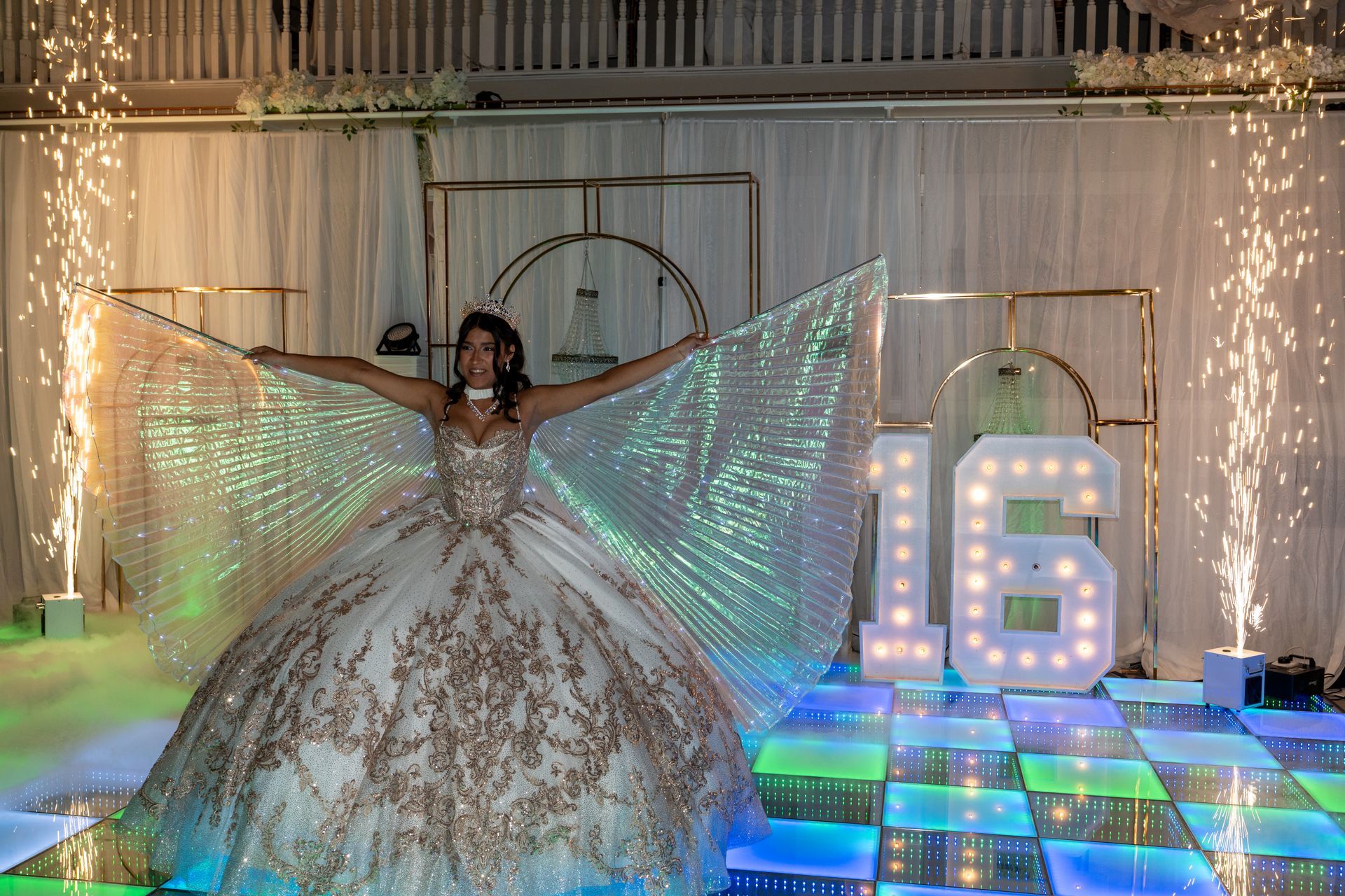 Teenager in a gown with sparkling wings, arms outstretched, celebrating 16th birthday. Fireworks, light-up numbers, dance floor.