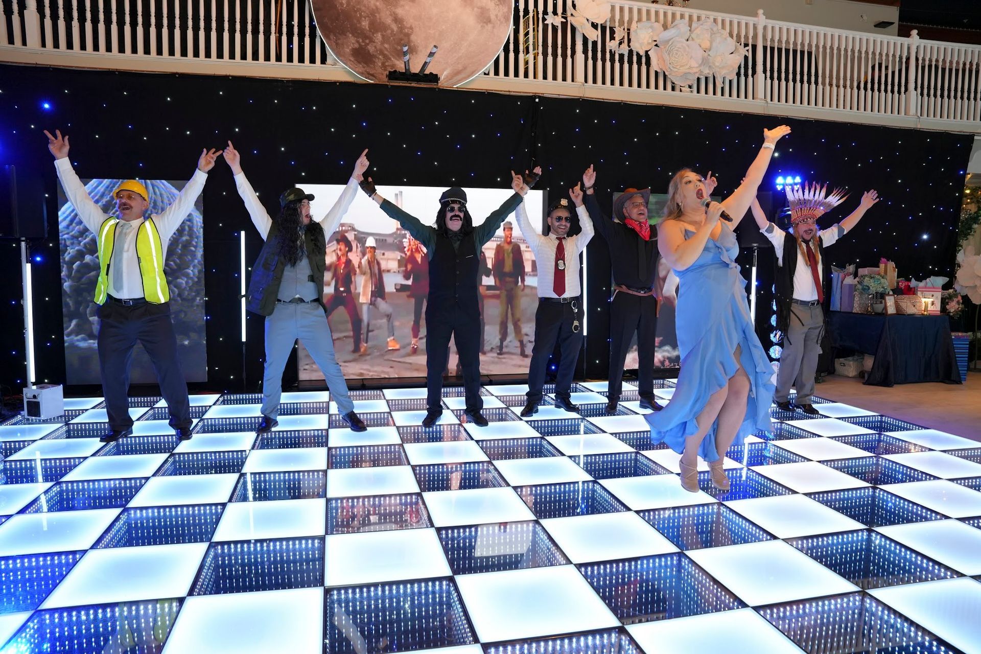 A woman sings on a checkered dance floor with costumed performers, arms raised, against a backdrop of stars and a moon.