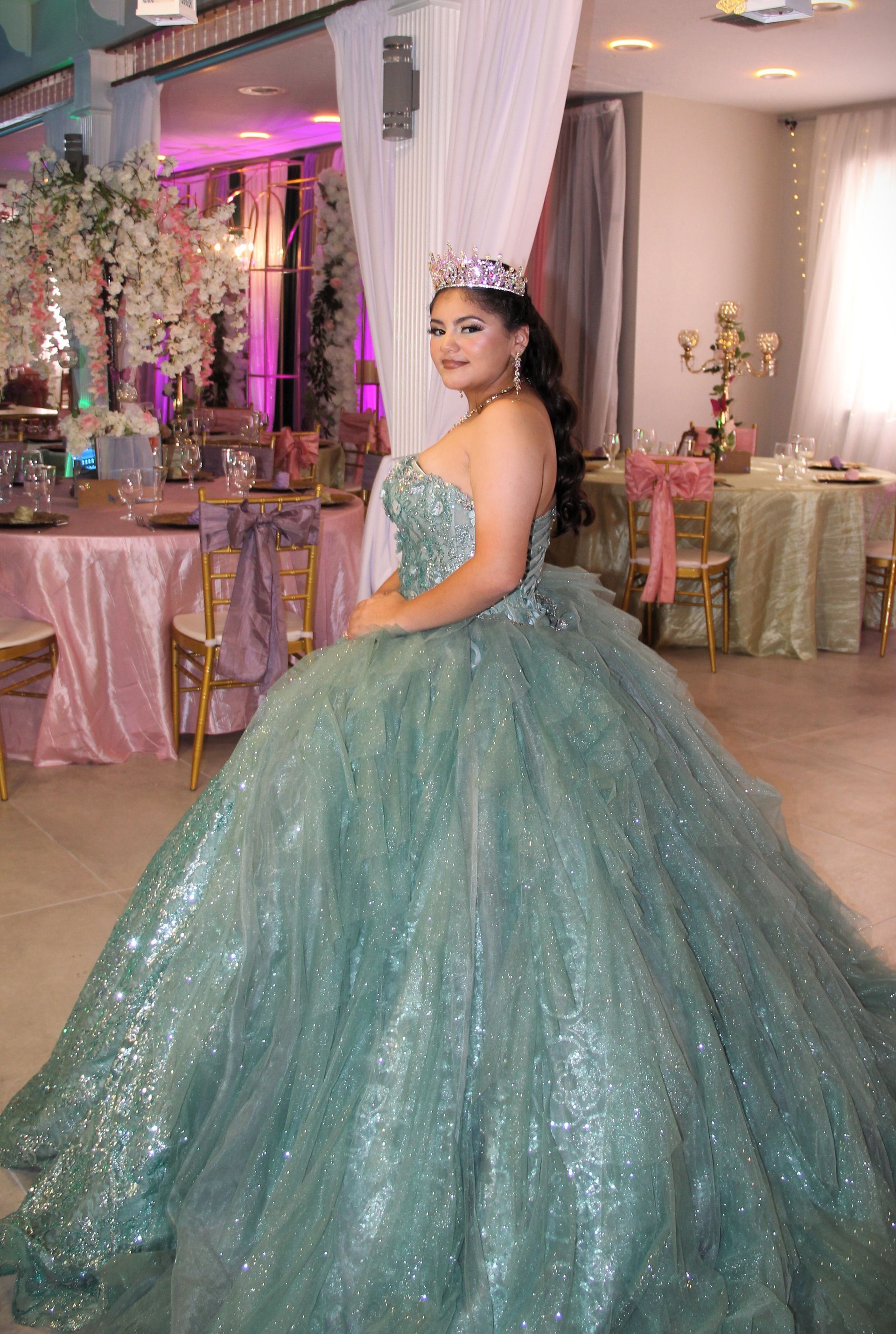 Woman in a sparkly, teal ballgown and tiara at a formal event, likely a quinceañera.