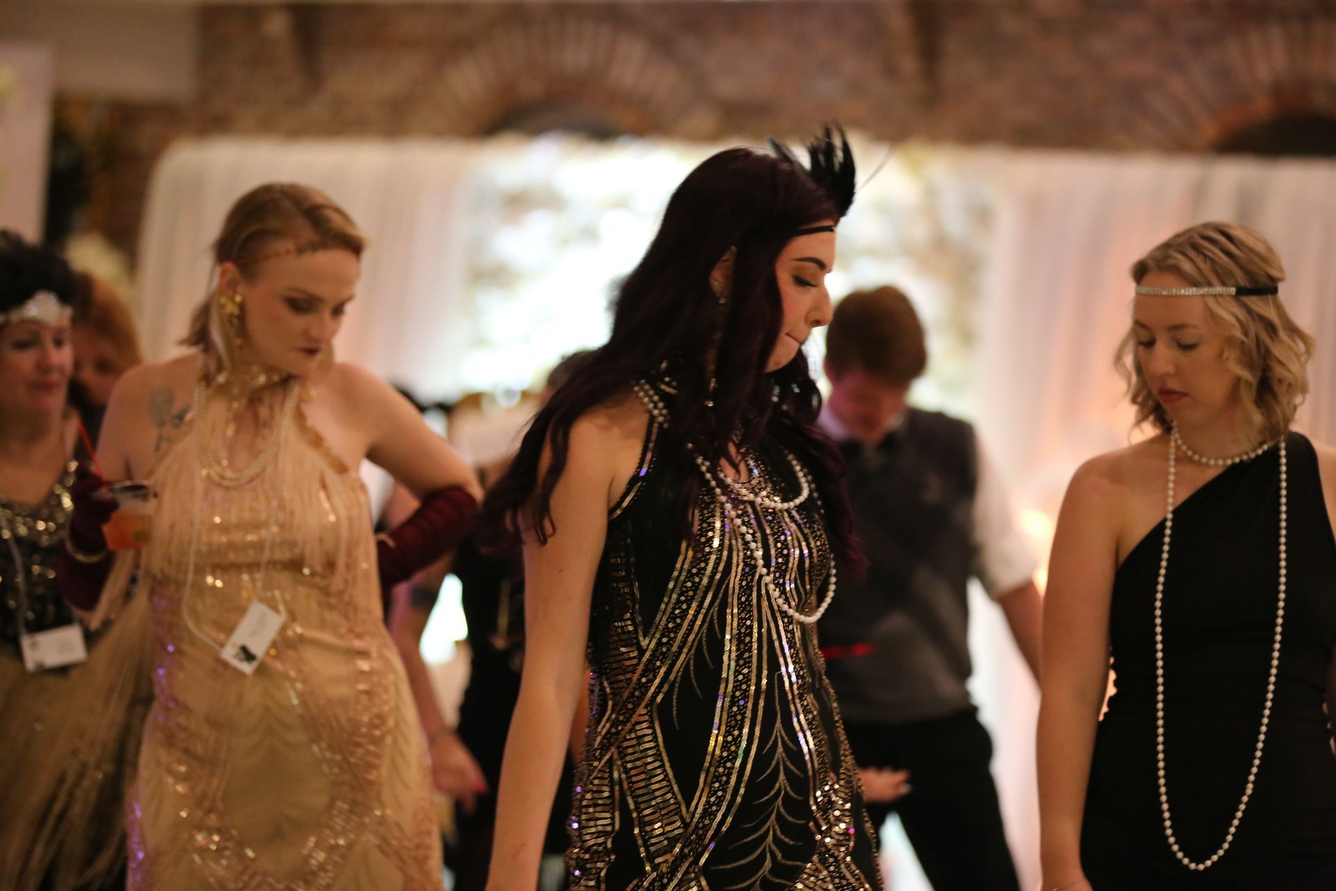 People in 1920s attire dancing in a decorated hall; woman in a black and gold dress in focus.