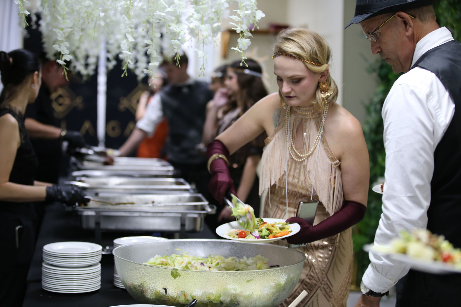 People at a buffet, one serving salad. Woman in flapper dress, others wear formal attire. Catering event with decorations.