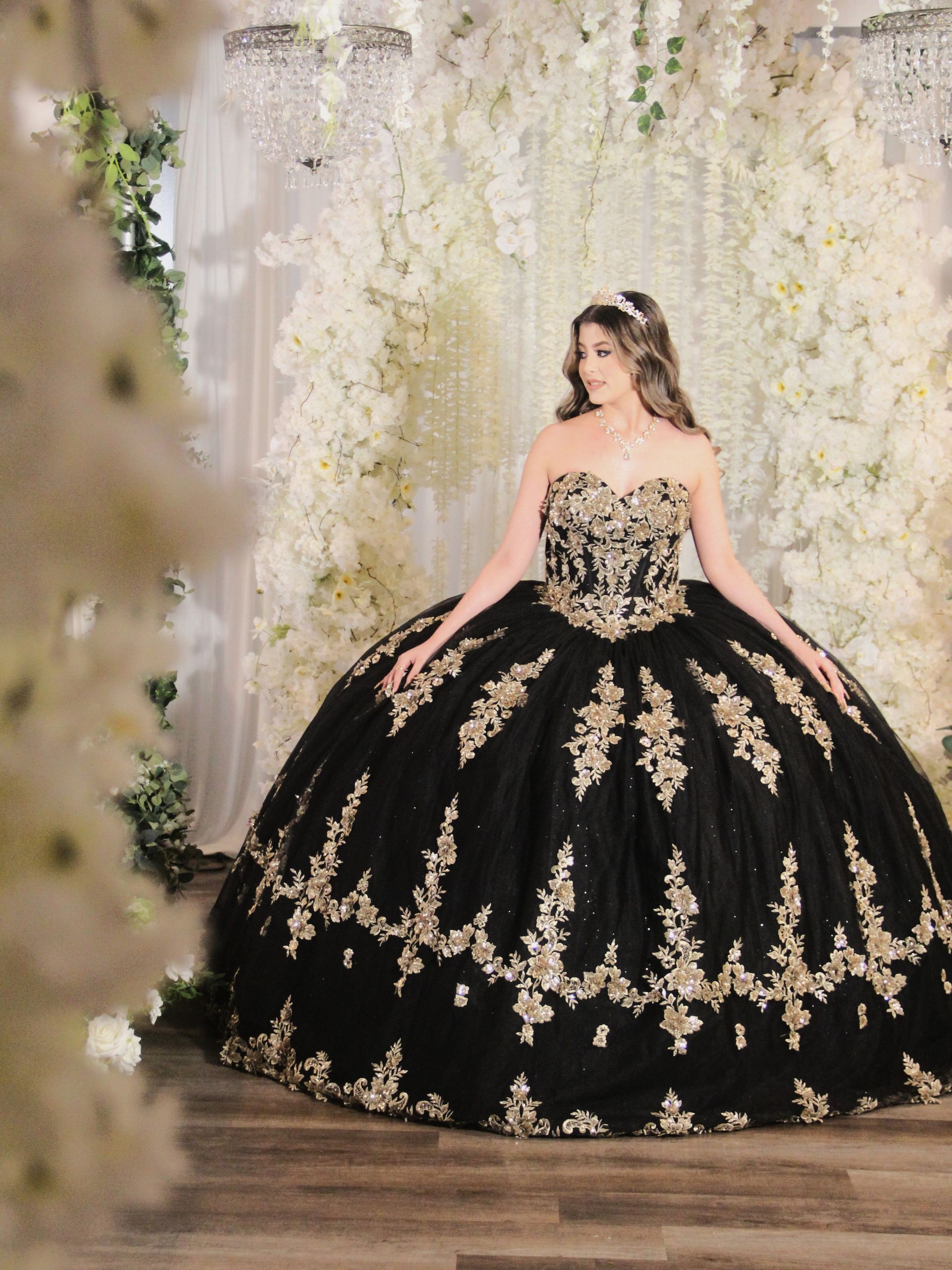 Woman in a black and gold ballgown poses against a white flower backdrop.