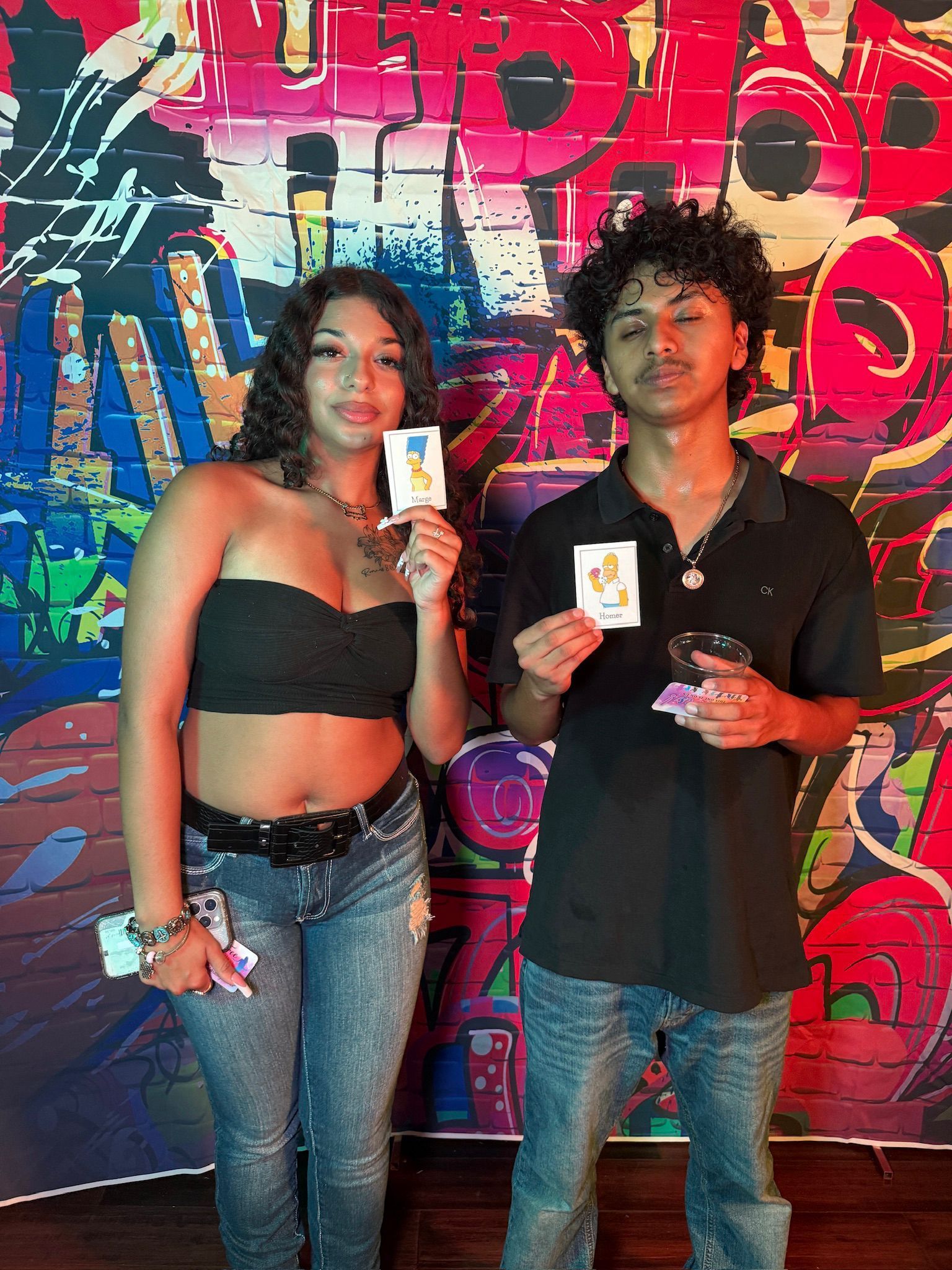 Two people holding cards, in front of graffiti wall. The person on the left wears a black top, jeans. The person on the right wears a black polo, jeans.