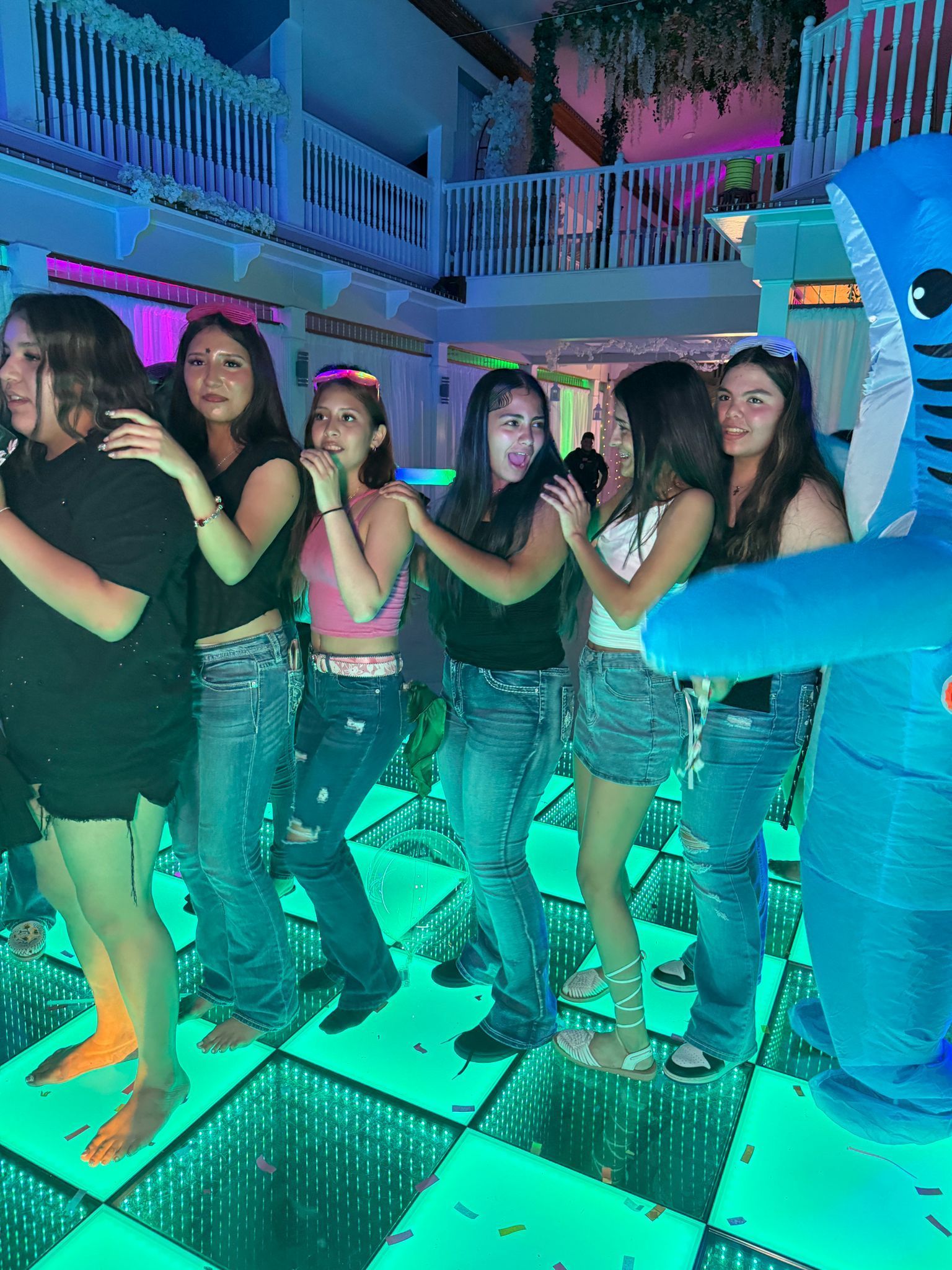 Seven young people linked together on a dance floor, some smiling, near a seahorse decoration.