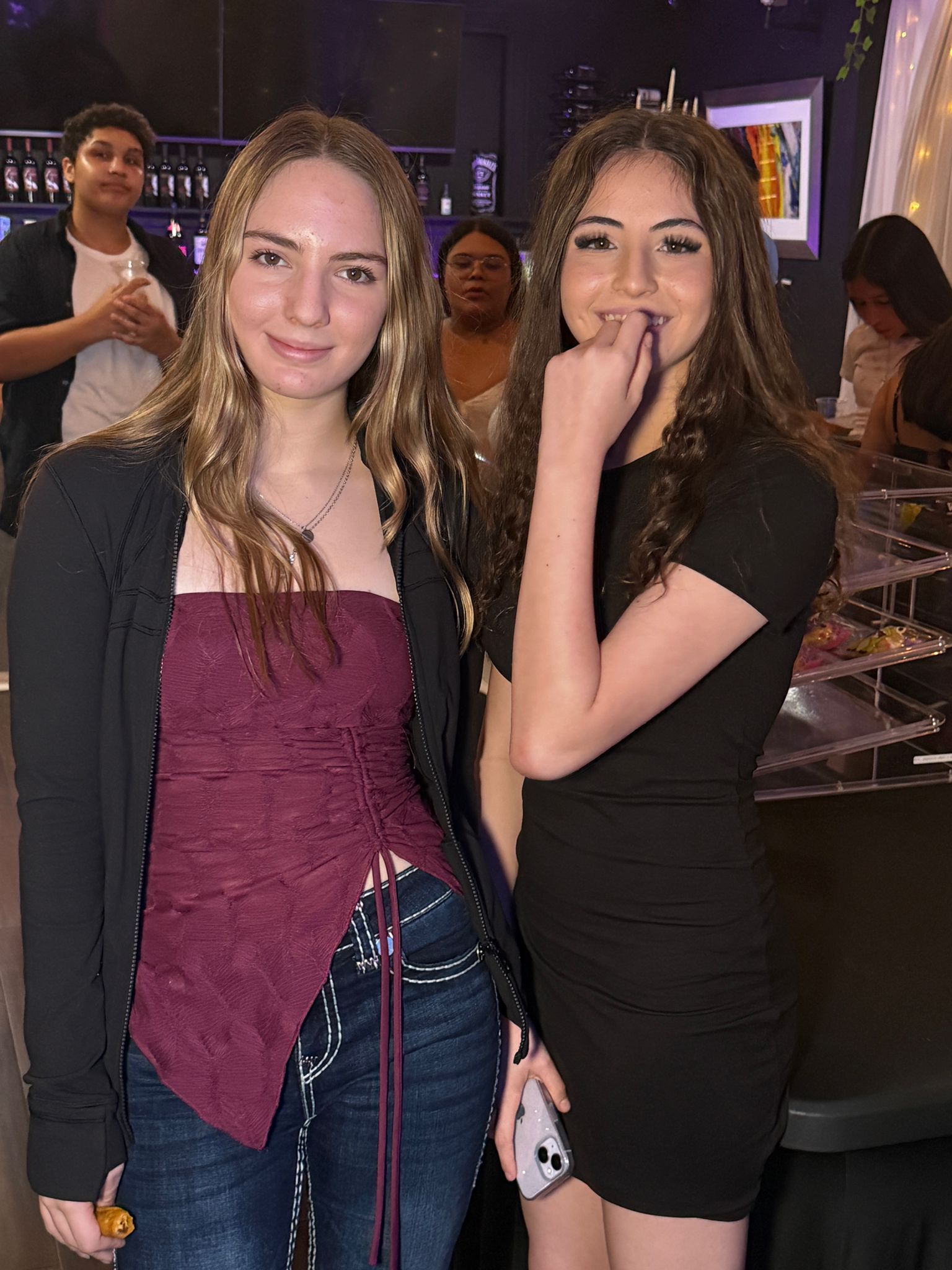 Two young women smile at a party; one in a maroon top, the other in a black dress, others in the background.