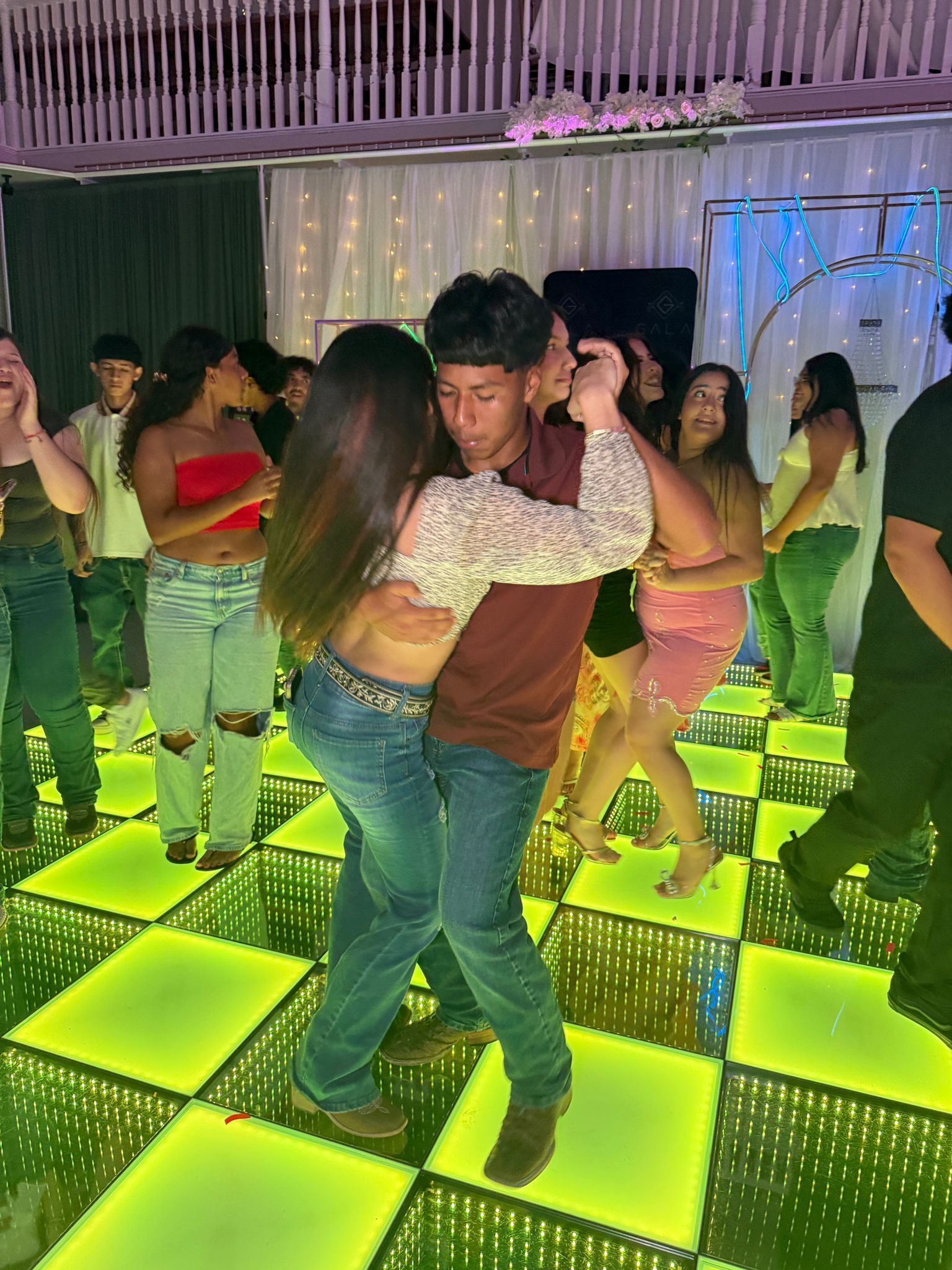 Couple dancing on a neon lit checkered dance floor, surrounded by other people in a party setting.