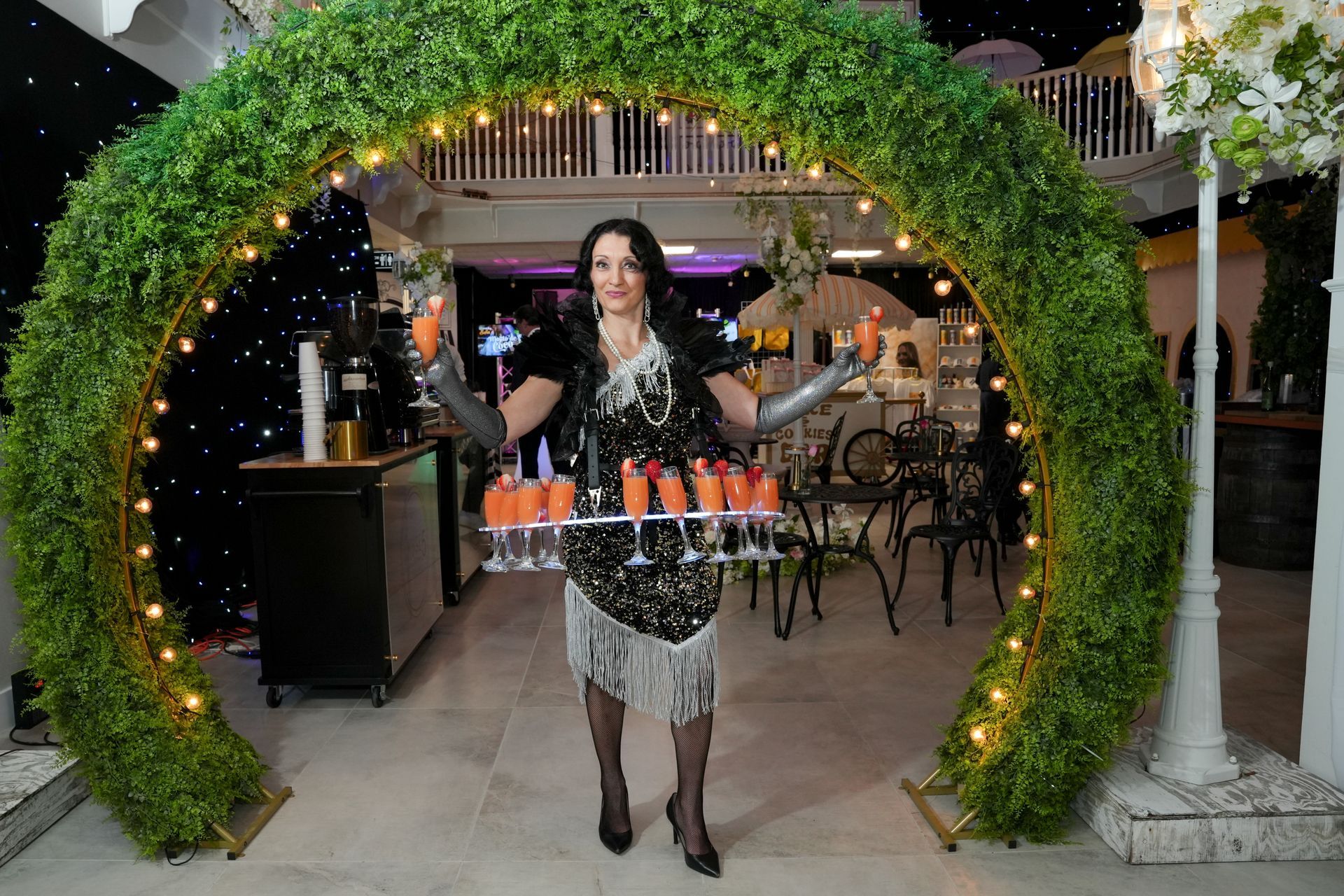 Woman in flapper dress carries tray of cocktails under a greenery arch with fairy lights.