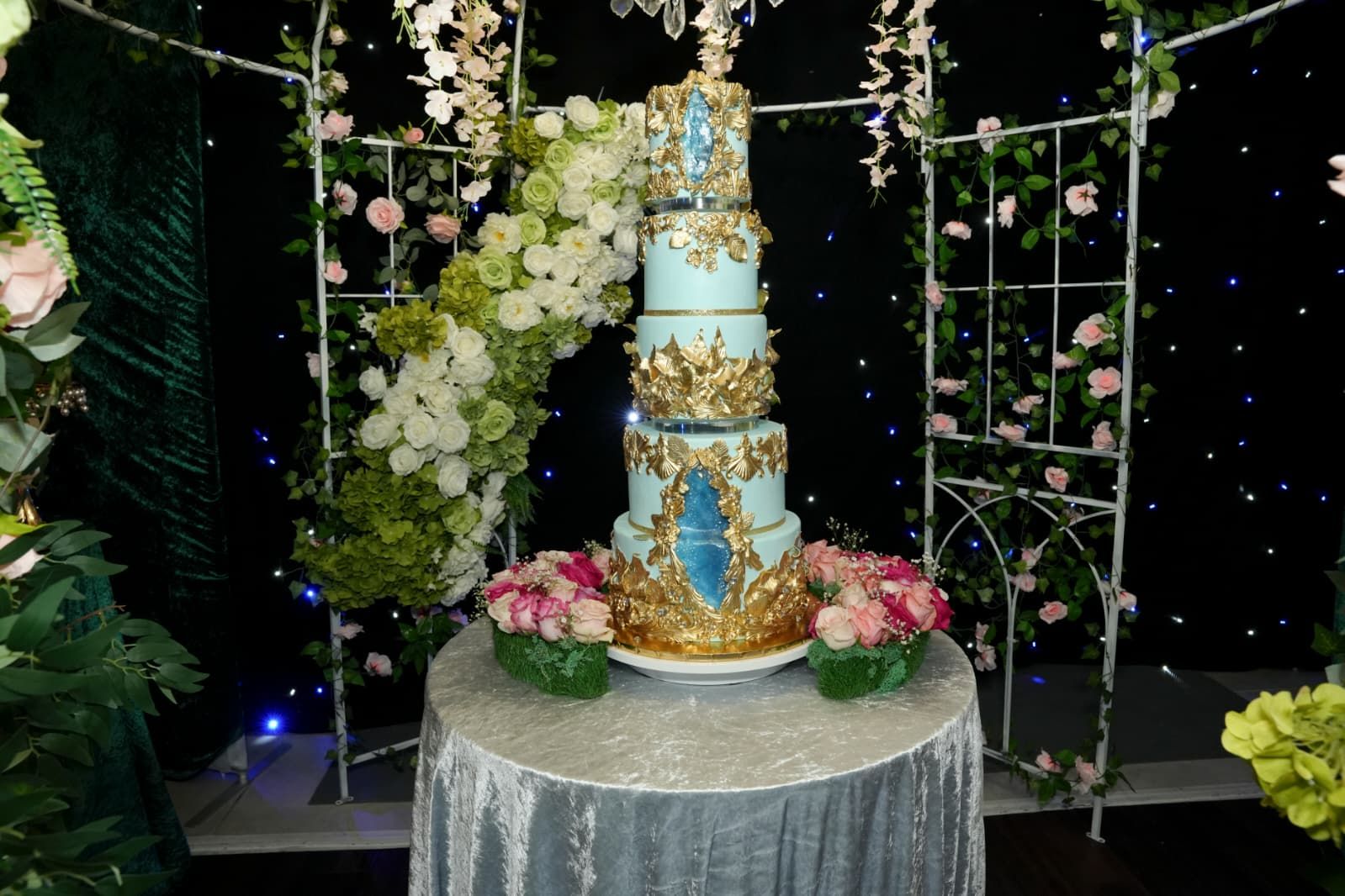 Elegant turquoise and gold tiered cake on a round table, adorned with flowers, in front of an archway.