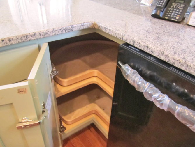 Corner kitchen cabinet with pull-out shelves, open. Light green door, beige countertop, and black appliance visible.