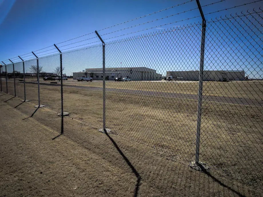 Chain-link fence beside a dry field under a clear blue sky, with industrial buildings in the distance.