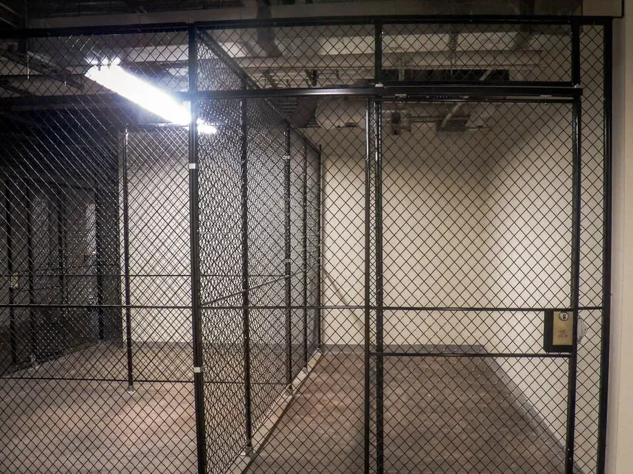 Empty chain-link fenced indoor enclosure with concrete floor and fluorescent lighting