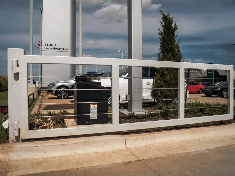 Modern white metal gate in front of a parking lot and office sign, with cars behind it.