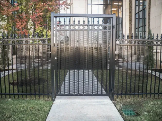 Black metal gate with arched top across a sidewalk, flanked by fence and building entrance.