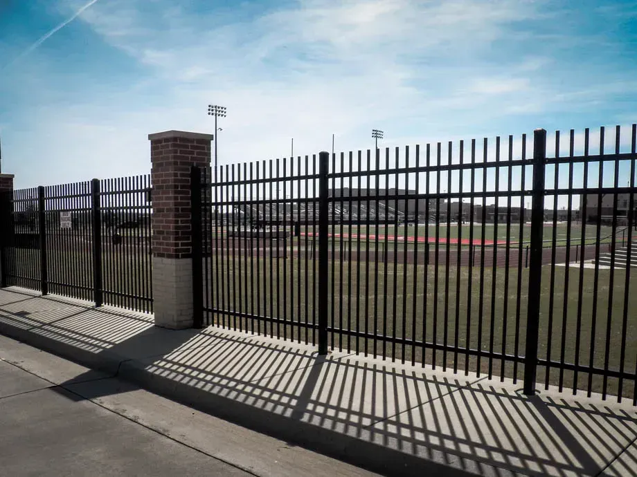 Black metal fence and brick gate beside a paved sidewalk, with a sports field behind it under a blue sky