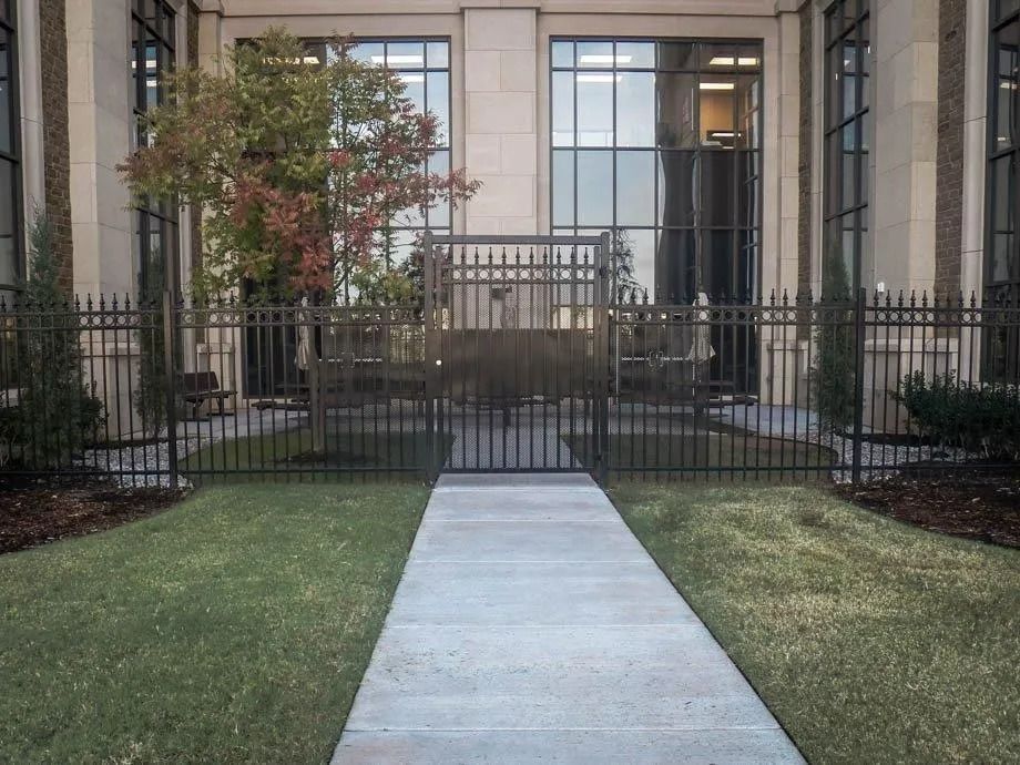 Concrete walkway leading to a gated entrance with steps, black iron fencing, and greenery outside a building