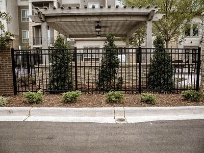 Apartment building entrance behind black metal fence and gate, with trees and sidewalk in front