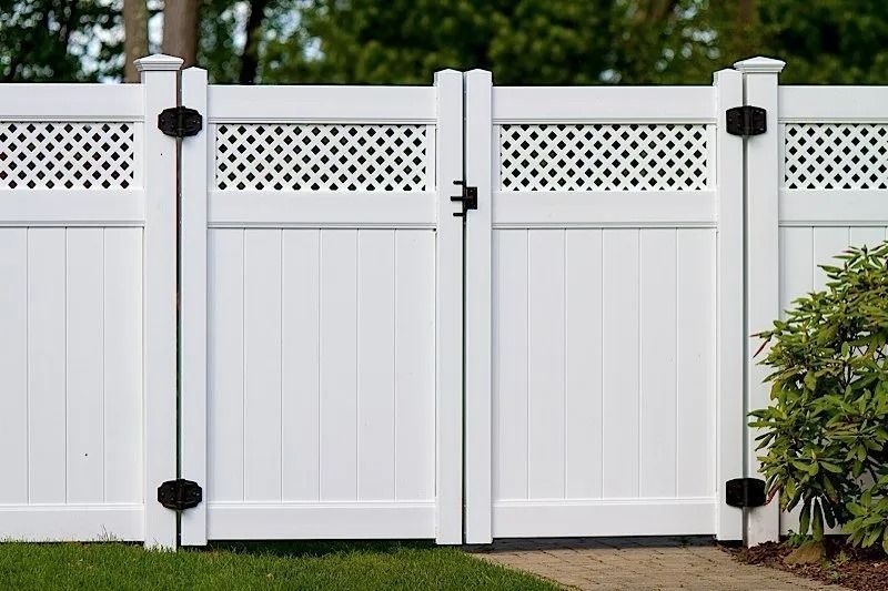 White vinyl privacy fence with lattice panels and black hinges along a grassy yard, with a shrub at right