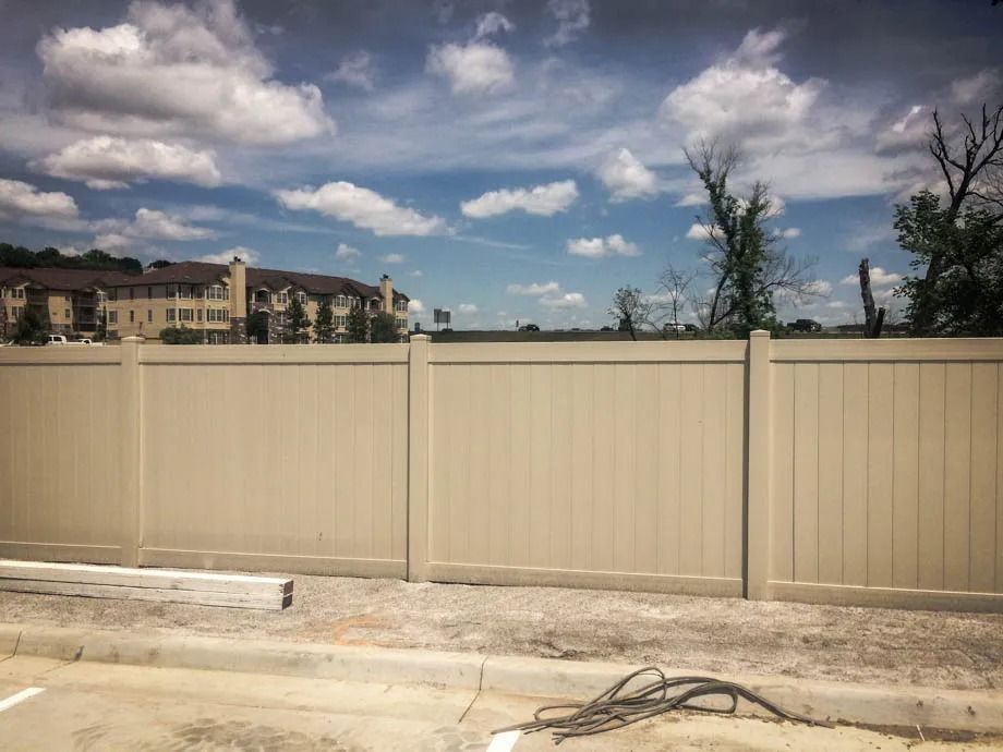 Beige fence along a street under a partly cloudy sky, with buildings in the background.