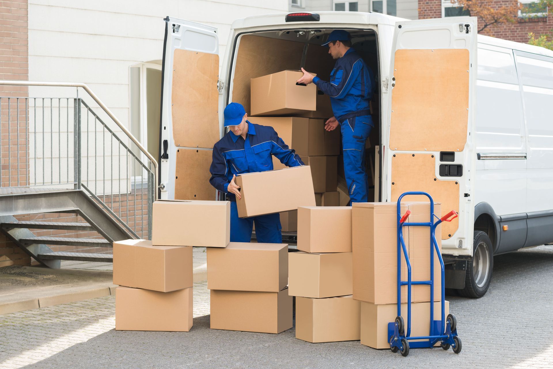 Two movers in blue uniforms loading cardboard boxes into a white van; a dolly sits nearby.