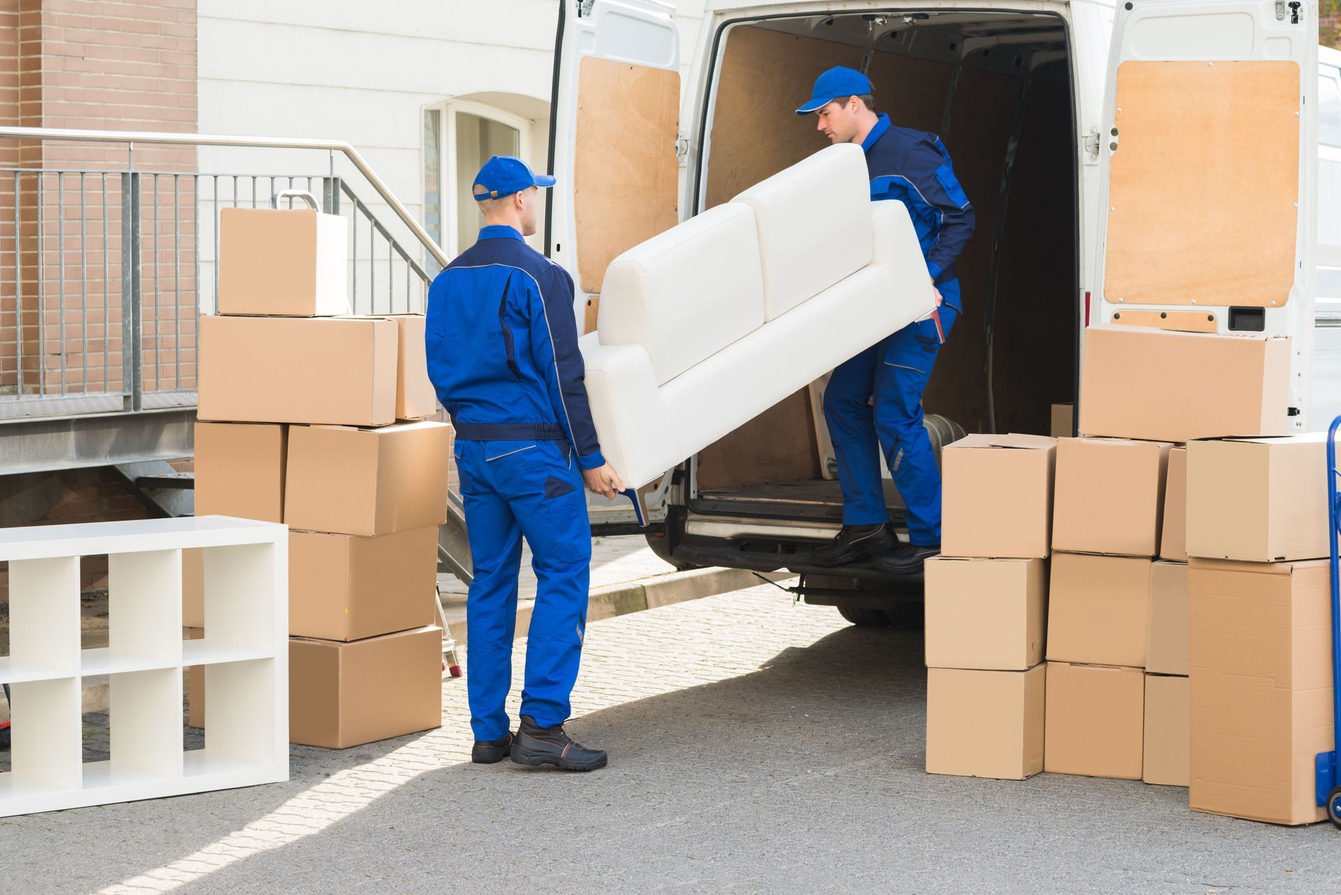 Two movers in blue uniforms loading a white couch into a white van, boxes stacked nearby.