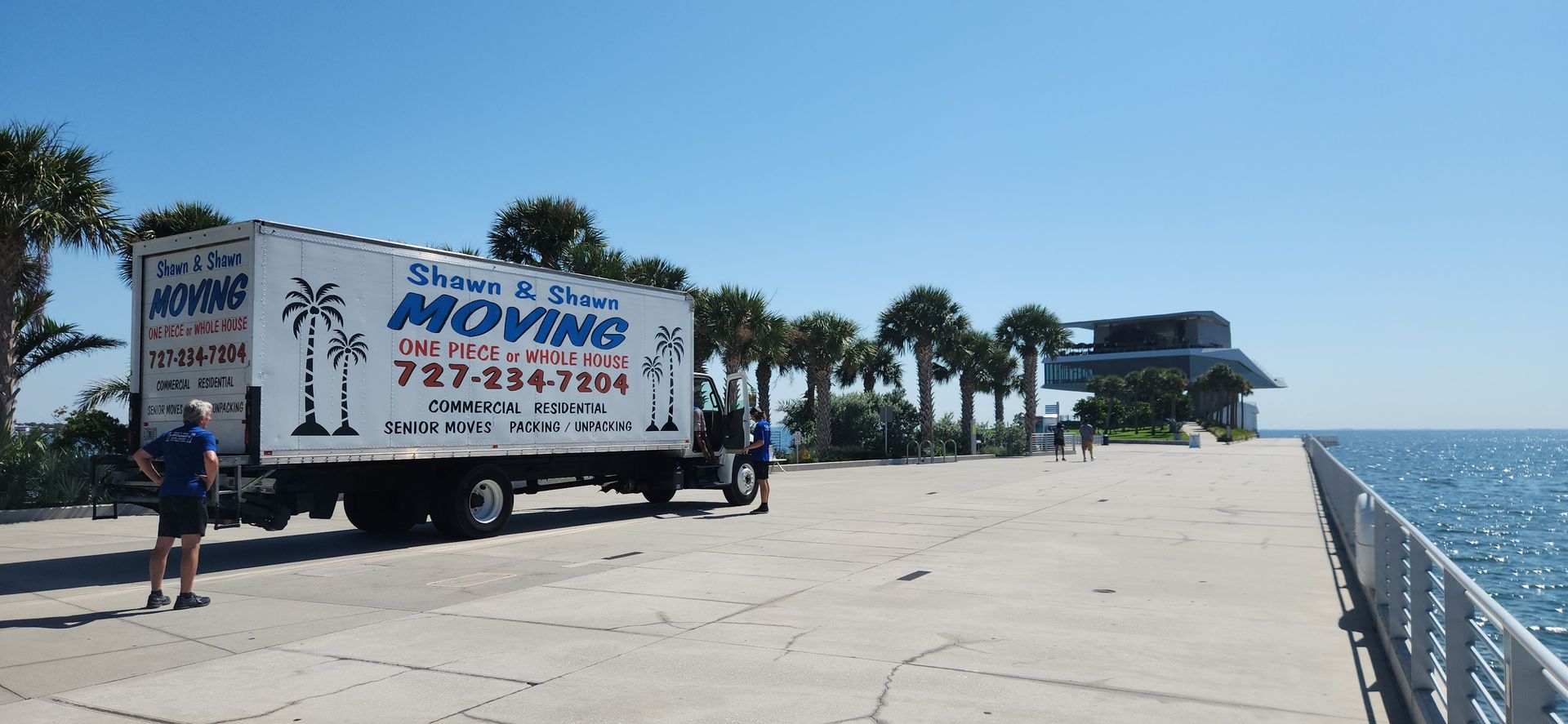 A moving truck parked on a waterfront walkway on a sunny day. Blue water and sky in the background.
