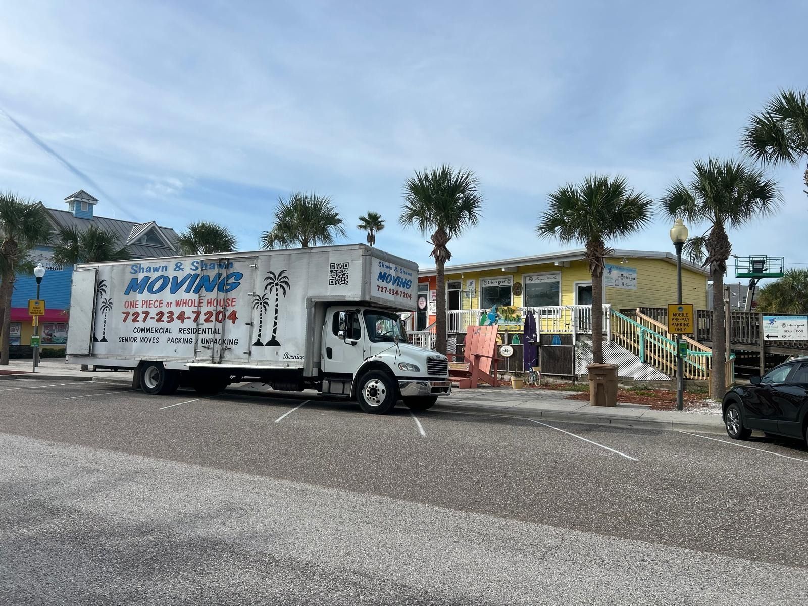Moving truck parked on a street near a yellow building with palm trees.