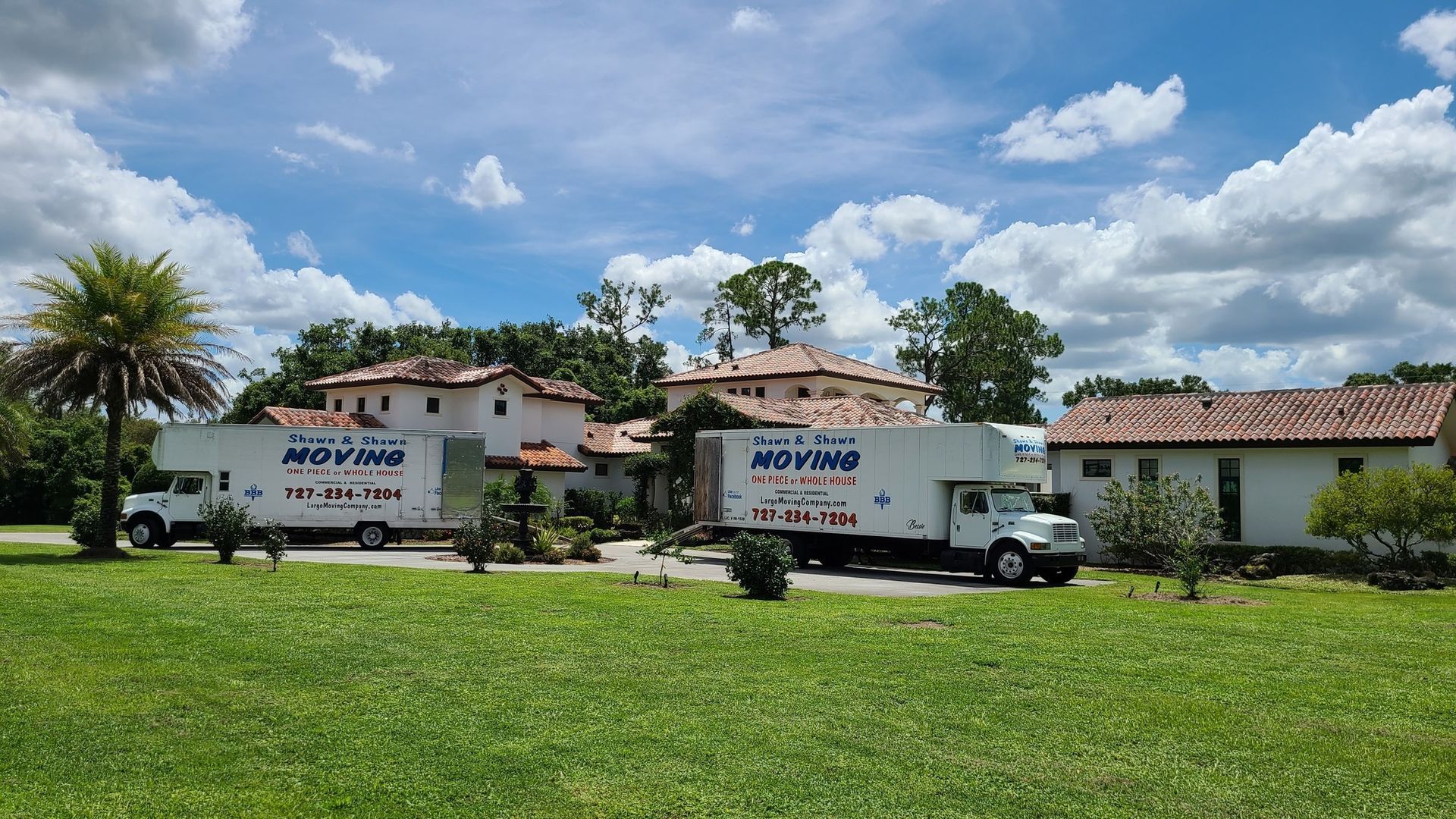 Two moving trucks parked in front of a large house with a green lawn on a sunny day.