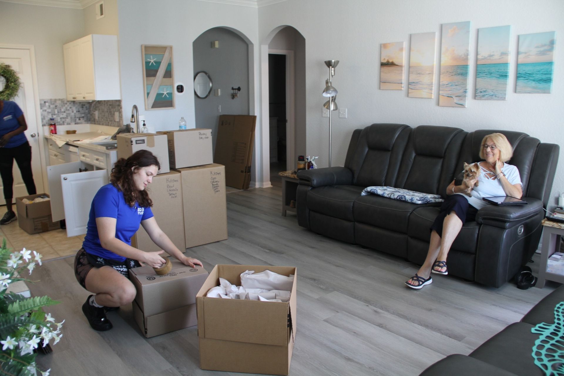 Movers unpacking boxes in a living room, woman watches from a black recliner.