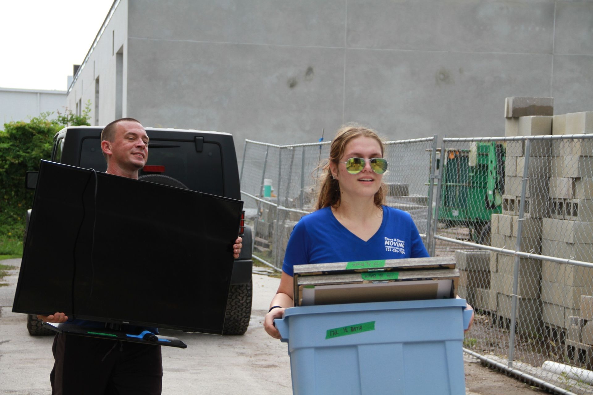 A man and woman carrying items outdoors. The man holds a black panel. The woman carries a blue bin.