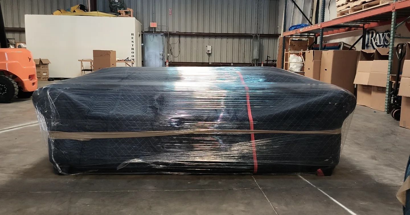 Large, black object wrapped in clear plastic in a warehouse. Orange forklift visible on the left.
