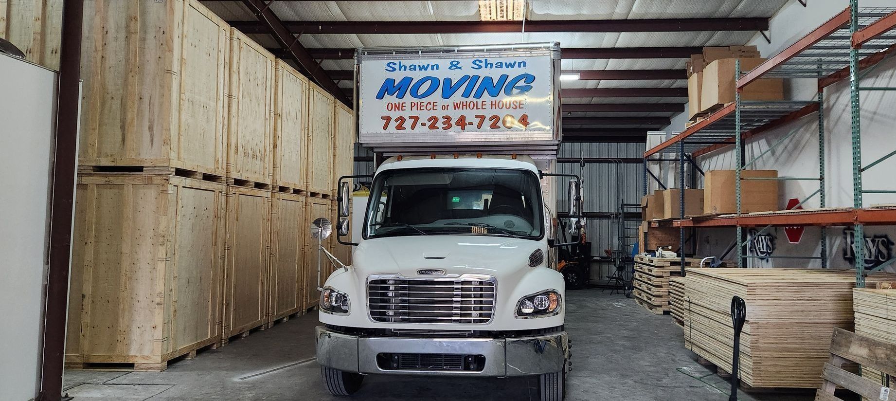 A white moving truck inside a warehouse, surrounded by wooden crates and storage shelves.