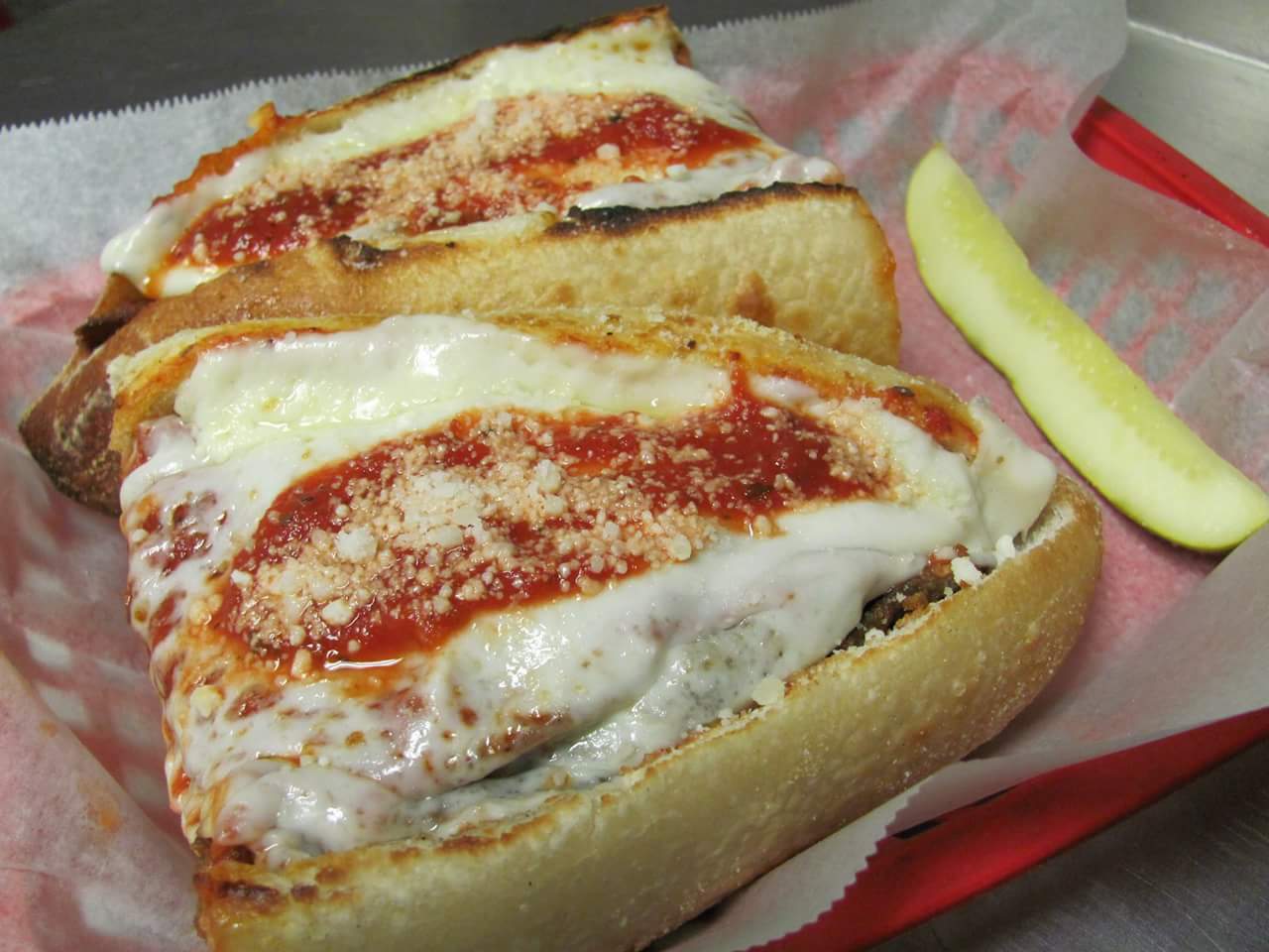 Two sub sandwiches with cheese and tomato sauce on a red tray with a pickle.