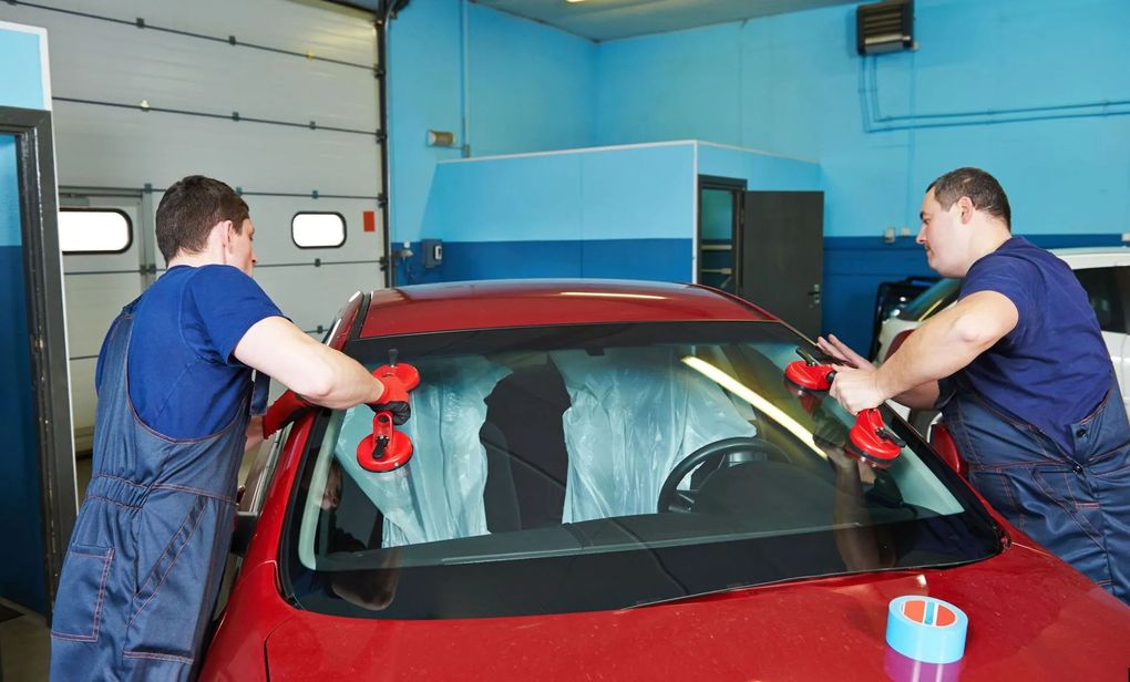 Two workers in blue shirts inspect a red car windshield in a garage using suction cups.